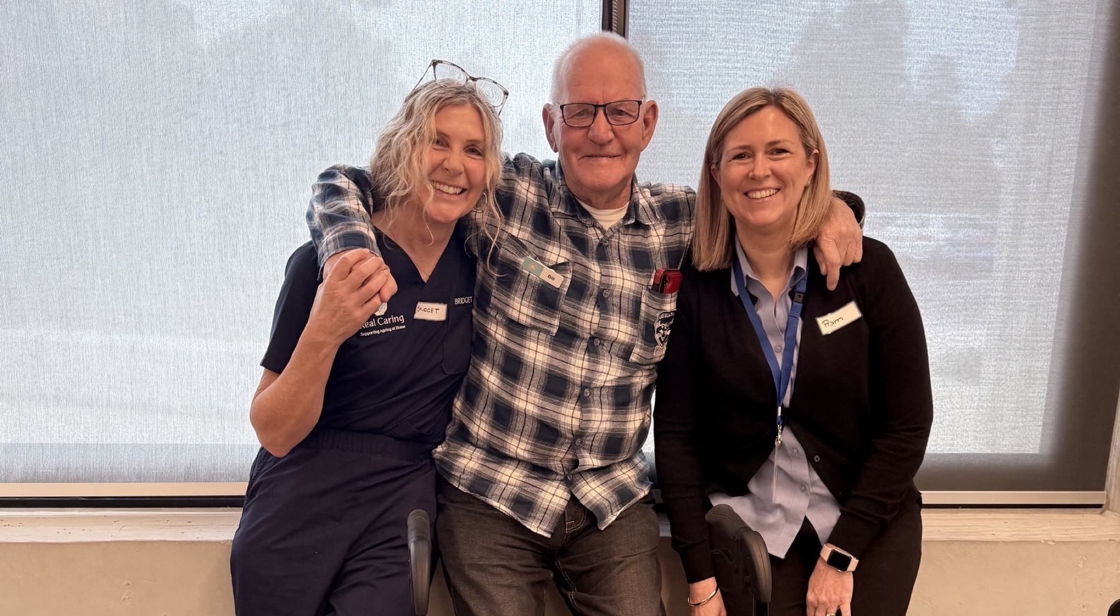 Bridget and Pam from Real Caring with Bill, a client, arms around each other and laughing